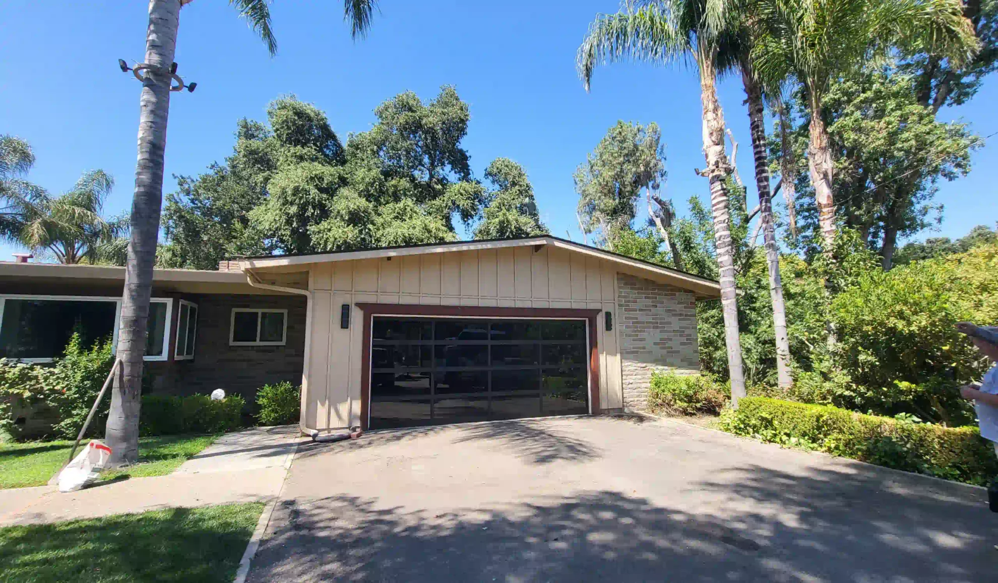 beige house with a brown roofing and a black garage door with some palms bushes and a parking outside